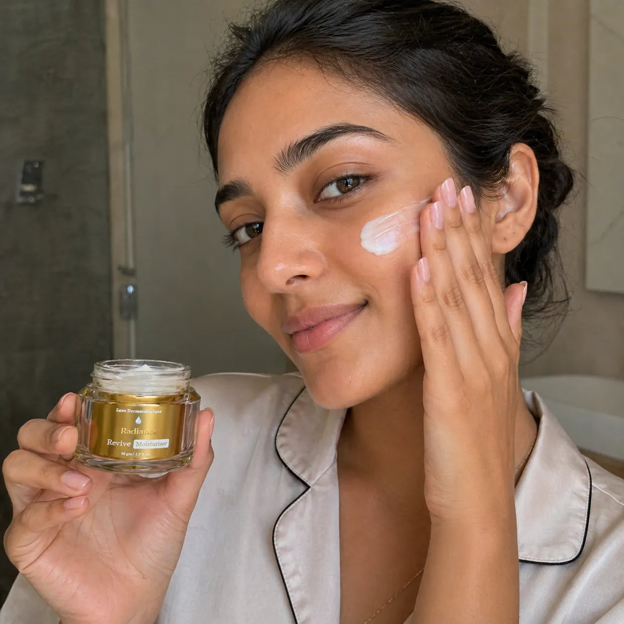 Woman applying cream to her face with a jar of cream in her hand, in a bathroom setting.